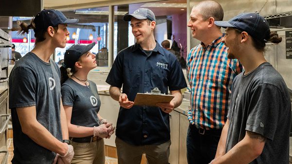 A Dewey's Pizza manager holding a clipboard meets with other employees. They gather in a circle and talk.