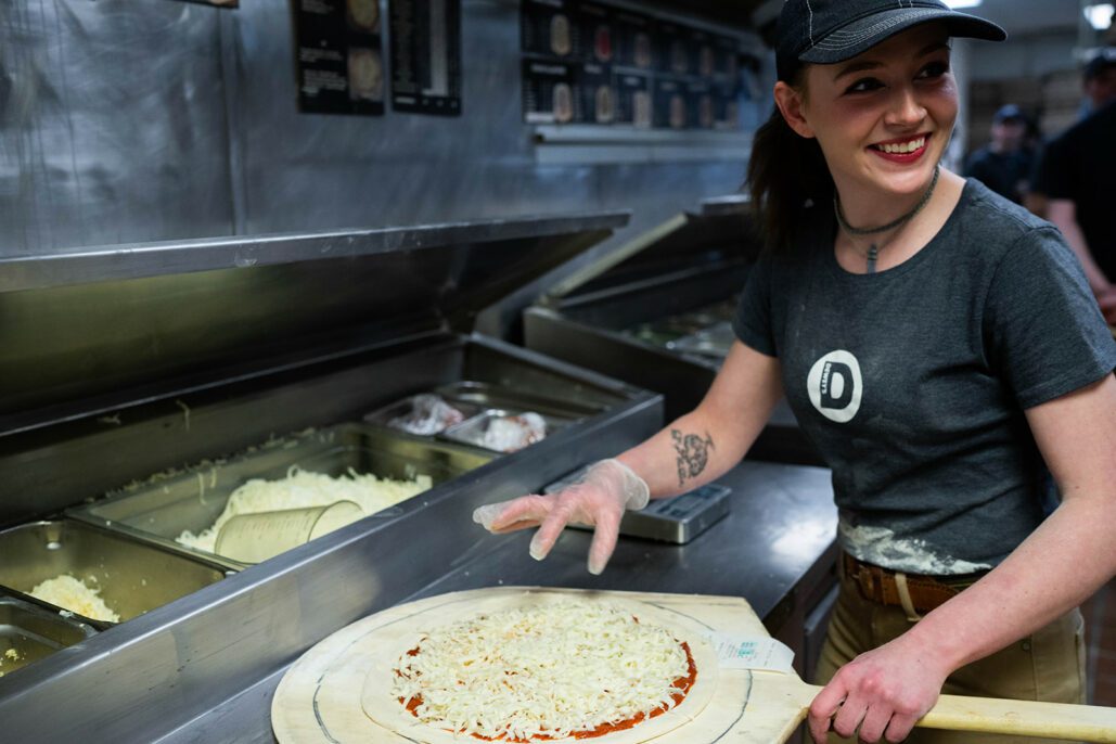 a dewey's employee adds cheese to a pizza