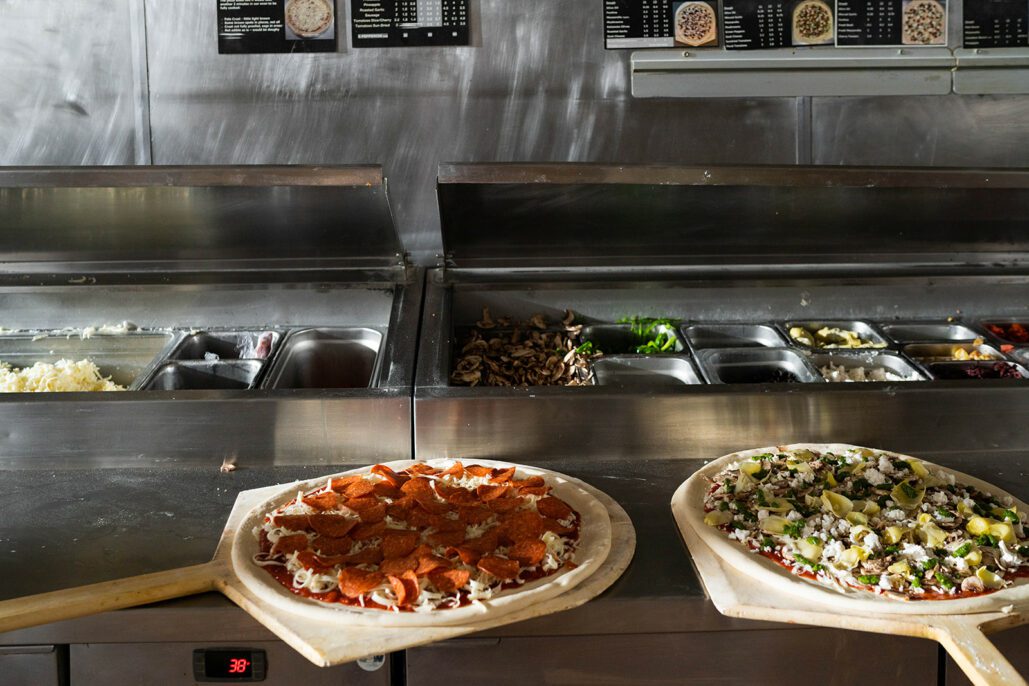 two uncooked pizzas sit near a toppings tray